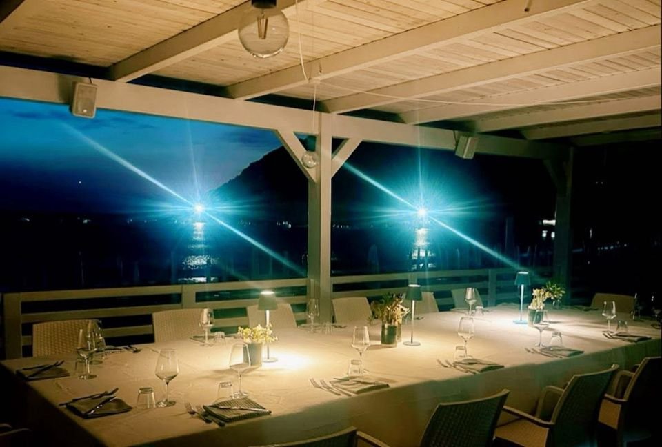 Lunga tavolata apparecchiata la sera sulla terrazza, con le lampade accese e il mare illuminato